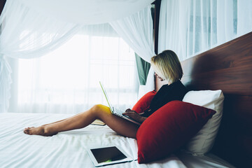 Skilled woman freelancer working on laptop computer sitting on bed at comfortable home interior,...