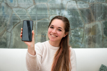 Mujer sonriente mostrando su celular con la pantalla apagada. Fotografía con enfoque selectivo en...
