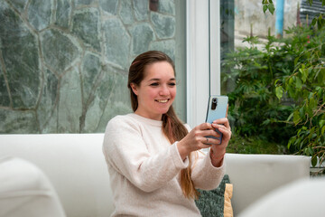 Mujer sonriente interactuando con su telefono movil sentada en su sofá de la sala de estar