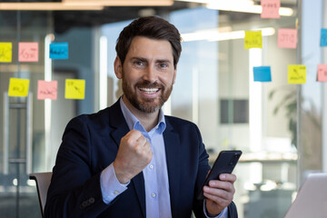 Mature boss businessman celebrating victory success, man at workplace with phone in hands, got good...
