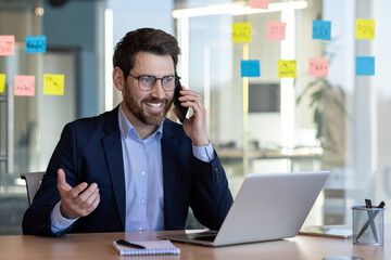 A man in a suit is talking on his cell phone while sitting at a desk with a laptop