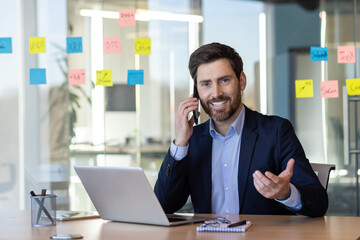 Obraz premium A man in a suit is talking on his cell phone while sitting at a desk. The scene suggests a professional setting, possibly in an office or a conference room. The man is focused on his conversation