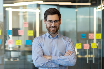 A man with glasses and a beard is smiling in front of a wall covered in sticky notes. The notes are...