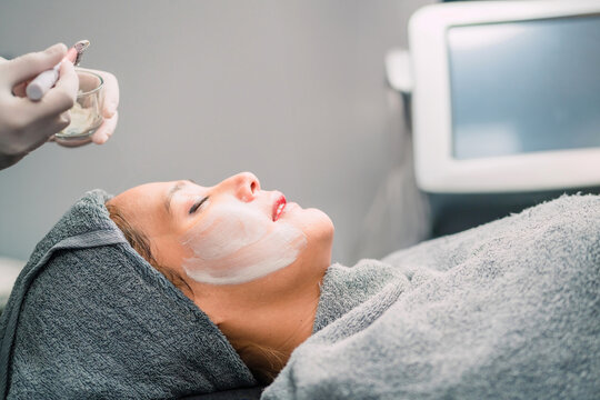 medical person brushing peeling woman face with cream. beauty doctor using cream rubbing on sleep patient using brush. asian patient relax in beauty salon got peeling cream spreading by gentle brush