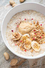 Oatmeal porridge served with peanut butter, banana slices and granola in a white bowl, vertical shot, middle close-up, selective focus