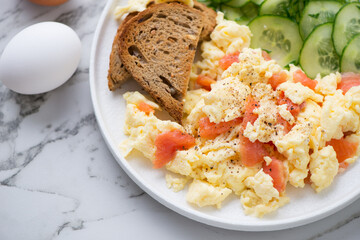 Scrambled eggs with smoked salmon, toasts and cucumber salad, horizontal shot on a white marble surface, middle close-up