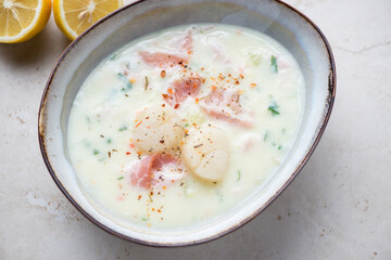 Bowl of milky chowder with scallops and bacon, horizontal shot on a light-beige stone background, elevated view