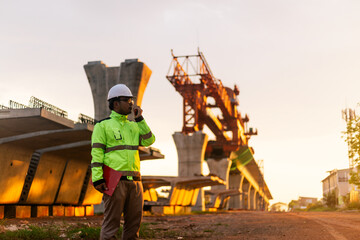 A construction worker is standing on a bridge, talking on his cell phone. The scene is set in a construction site, with a large crane in the background. The worker is wearing a yellow jacket