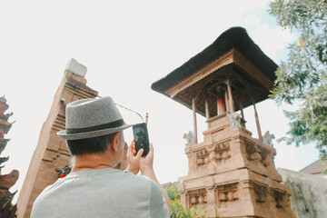 An adult man with grey hat is capturing photo of traditional Balinese Hindu temple with bokeh or...