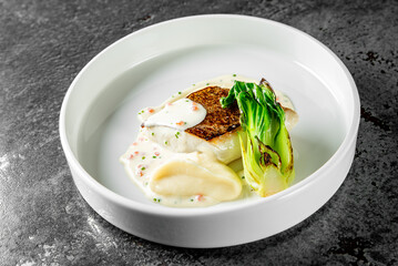 An elegant gourmet dish with grilled fish and seared bok choy, served with a creamy sauce in a white bowl on a textured grey surface