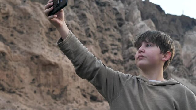 An 11-year-old teenager with a lot of hair and a fluffy hairstyle takes pictures of himself in the mountains using a mobile phone camera with a smartphone at arm's length. Selfie at sunset