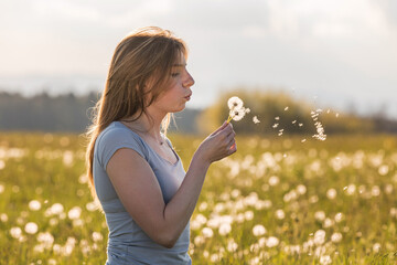 Young woman blowing dandelions, on the green meadow with the summer sun in the background. Dream, and life optimism concepts.