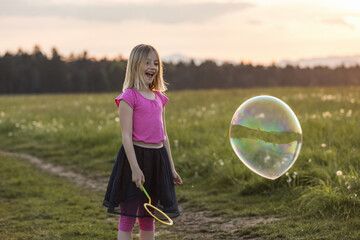 Smiling little girl playing with bubble wand making big bubbles on the green meadow for summer fun. Outdoors play activities concept.