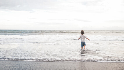 A happy toddler boy is running joyfully into the ocean water on the beach, under the blue sky and fluffy clouds. The liquid and fluid waves splash around him as the wind gently blows. Mothers day.