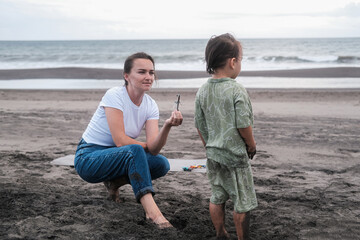 Mom and son in casual clothes play while sitting on the beach on black sand near the ocean. A young woman shows a toy airplane to a little boy and smiles. Mothers Day. Summer holidays with your child.