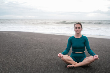 Fototapeta na wymiar A young woman in lotus position sits on a black sand beach and meditates against the backdrop of the ocean with big waves. Practice mindfulness and purity of mind. Concentration. Summer sports.