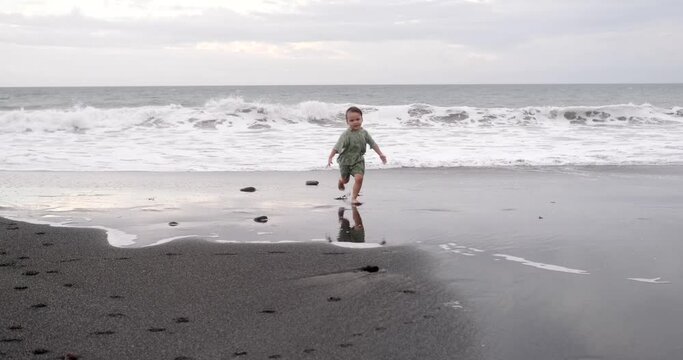 Happy little boy running on a black sand beach near the ocean and fooling around. He plays around and flaps his arms like a bird. Summer holiday with the whole family. Mothers Day.