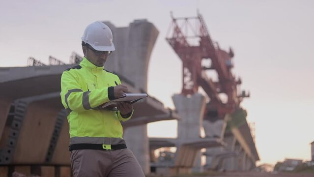 A construction worker is standing on a bridge, talking on his cell phone. The scene is set in a construction site, with a large crane in the background. The worker is wearing a yellow jacket