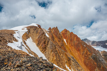 Snow-capped mountain ridge with cornice and red sharp rocks on top in sunlight. Snow cap and shiny...