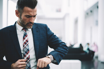 Confident mature entrepreneur dressed in formal wear checking time on watch standing in office of...