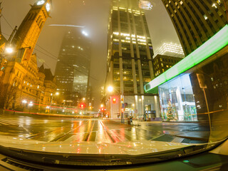 Fototapeta premium Long exposure pov of car driving through city of Toronto downtown. Ontario, Canada. Travel trip, dash cam or car camera view forward at night rainy city.