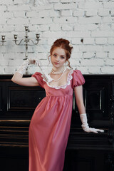 Young red haired woman in empire style dress, high gloves and fan standing near piano. Historical...