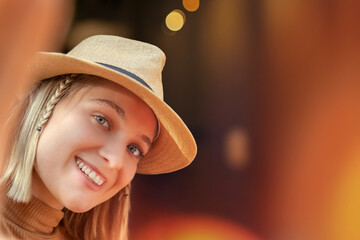 Young happy girl with hat and smiling at the camera, taking selfie outside with orange lights bokeh background - Youth concept
