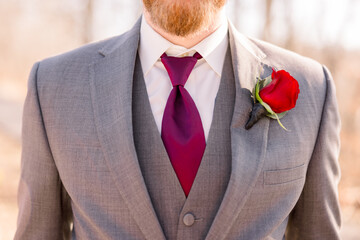 A groom wearing a gray suit and maroon tie and a rose boutonniere. 