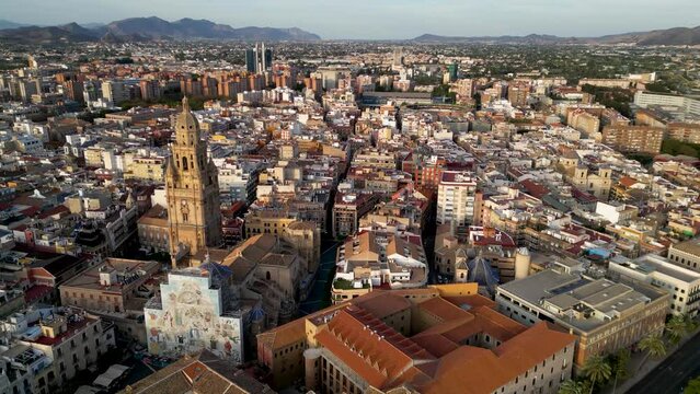 Aerial view of Murcia. Drone slowly circling above the city center. Cathedral of Murcia and Cardenal Belluga Square. Sunset warm colours. Mountains in background. Travel
