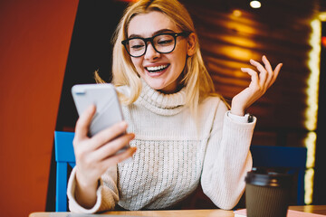 Obraz premium Cheerful hipster girl excited with good news reading mail on smartphone sitting in coffee shop, emotional woman happy about getting discount for internet communication checking news on telephone