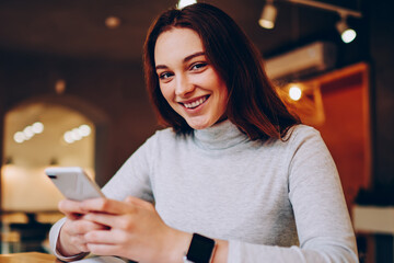Portrait of cheerful hipster girl using smartphone for chatting having fun adding  friends in social networks  on coffee break, happy young woman looking at camera holding cellular connected to wifi