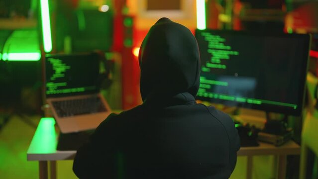 Hacker writing code cracking computer. Man programmer in hoodie cracks system, using computer in dark room back view. Hacker attack, technology, virus software, digital, cyber security concept.