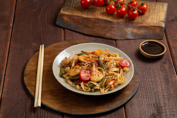 udon shrimp in pad thai sauce on a wooden table next to cherry tomatoes, soy sauce and chopsticks