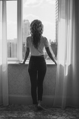 The girl stands at full height by the window with her back to the camera
