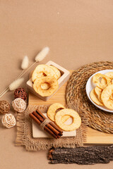 Crispy apple chips in a white round plate and in a wooden box, cinnamon sticks on a wooden desk on a beige background in rustic style. Healthy sweet snack.