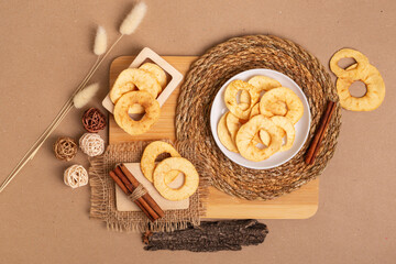 Crispy apple chips in a white round plate and in a wooden box, cinnamon sticks on a wooden desk from above on a beige background in rustic style. Healthy sweet snack.