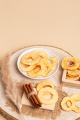 Tasty sweet apple chips in a plate and in a wooden box, cinnamon sticks on a wooden desk on a beige background.