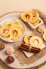 Tasty sweet apple chips in a plate and in a wooden box, cinnamon sticks on a wooden desk on a beige background.