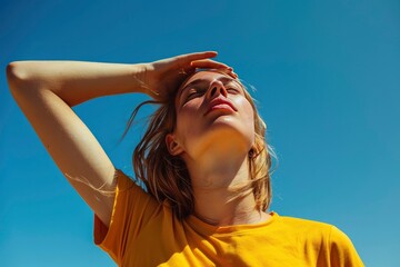 Young woman in a yellow t-shirt with her hand on her forehead trying to cool down from the heat and sun, under a blue sky.