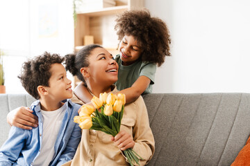 Excited young woman holding bunch of flowers while looking at her daughter, hugging son at home