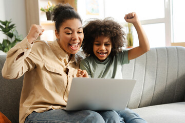 Happy mother and her daughter looking at laptop screen while holding hands up, celebrating at home