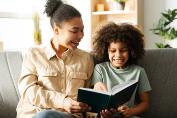 Happy young woman sitting next to her daughter on cozy sofa at home while reading book