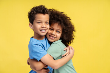 Cute brother and sister hugging, looking at camera, posing in studio, isolated on yellow background