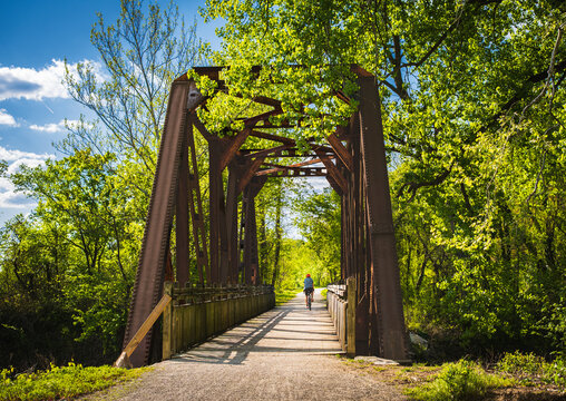 View of railroad bridge  converted into recreational trail in summer; male bicyclist riding  in background; lush greenery on both sides; blue sky