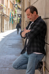 An elderly man leans against a wall, arms crossed on his chest, looking down, street portrait.
