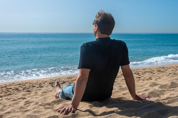 An elderly man sitting on a sandy beach looking out to sea, rear view.