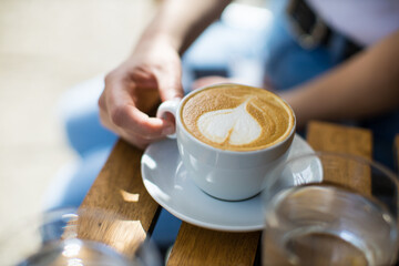 A cup of cappuccino with heart shaped foam being held by woman's hand on a wooden table
