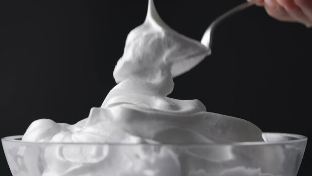 Chef takes whipped egg whites with sugar with a spoon from a glass bowl. Close-up of food on a black background