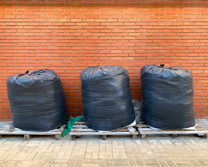 Three big black plastic garbage bags full with trashes placed in the street on pallets against the red brick wall.