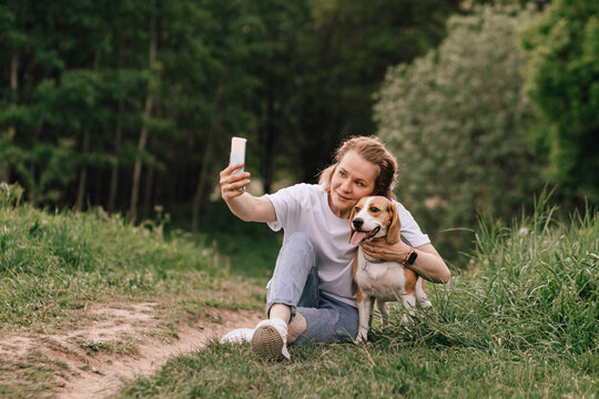 A woman takes a selfie cuddling her beagle dog while relaxing in the park on a summer day. Concept of social media, well-being, pet products - Powered by Adobe